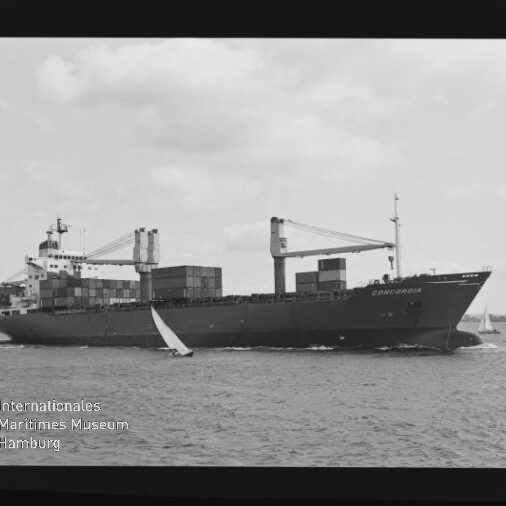 Schwarzweißaufnahme eines Containerschiffs namens „Concordia“ mit gestapelten Containerboxen und zwei Ladekränen, das auf dem Meer fährt und von kleinen Segelbooten begleitet wird; Filmrand mit Aufschrift AGFA APX 100 und Museumswasserzeichen unten links.