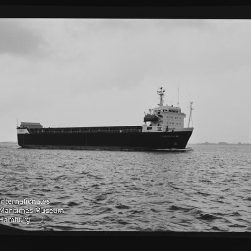 Schwarzweiß-Aufnahme eines Frachtschiffs in Seitenansicht auf ruhiger See vor bewölktem Himmel, im Vordergrund welliges Wasser und unten links ein Logo mit der Aufschrift „Internationales Maritimes Museum Hamburg“.