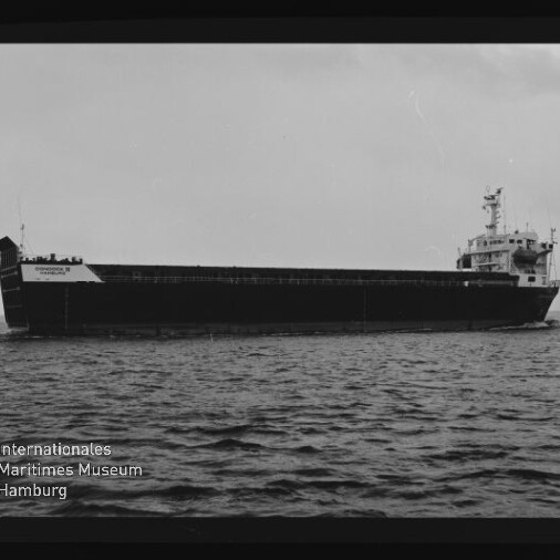 Schwarzweiß-Seitenansicht eines großen Massengutfrachters auf ruhiger See mit langem Ladebereich und achtern gelegenem Steuerhaus, unten links Wasserzeichen des Internationalen Maritimen Museums Hamburg.