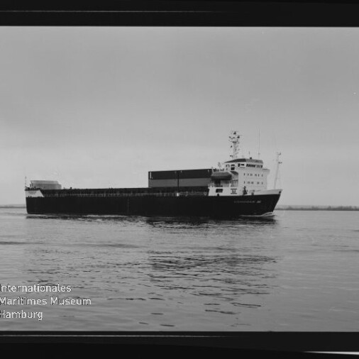 Schwarzweiß-Foto eines Frachtschiffs in Seitenansicht auf ruhigem Wasser, mit sichtbarem Aufbau, geladenen Deckseinheiten, weiter Horizont und dem Logo des Internationalen Maritimen Museums Hamburg unten links.