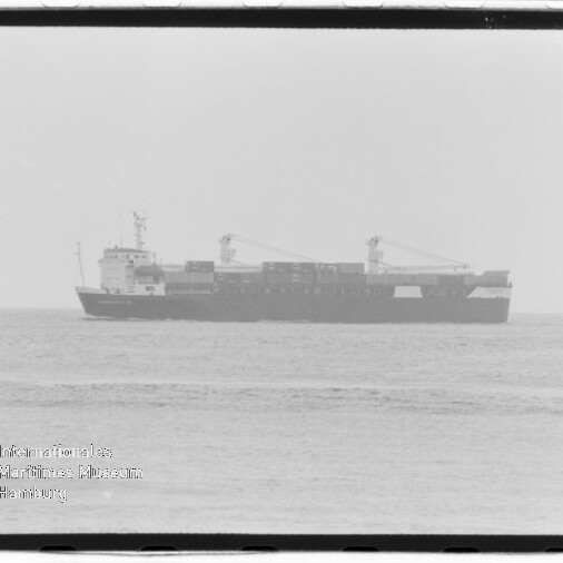 Schwarzweißaufnahme eines Containerschiffs im Profil auf ruhiger See, mit gestapelten Containern und zwei Ladekränen, unscharfer Horizont und Wasserzeichen des Internationalen Maritimen Museums Hamburg unten links.