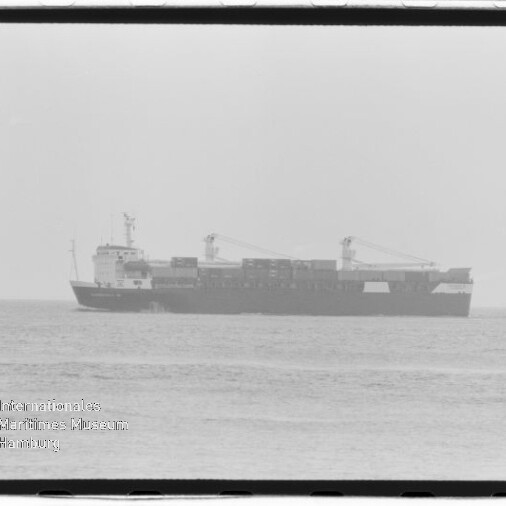 Schwarzweiß-Aufnahme eines Containerschiffs in Seitenansicht auf ruhigem Meer mit gestapelten Containern und zwei Ladekranen, unten links Wasserzeichen „Internationales Maritimes Museum Hamburg“ und sichtbarer Filmrand.