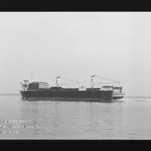 Schwarzweiß-Foto eines Frachtschiffs mit zwei Deckskränen und achterem Aufbauten, das ruhig auf dem Meer nahe der flachen Küstenlinie fährt; links unten sichtbar das Wasserzeichen des Internationalen Maritimen Museums Hamburg.