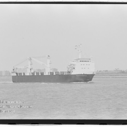 Schwarzweiß-Foto eines Frachtschiffs mit Aufbauten und zwei Ladekränen, das auf ruhigem Meer vor einer flachen Küstenlinie fährt; Wasserzeichen des Internationalen Maritimen Museums Hamburg unten links.