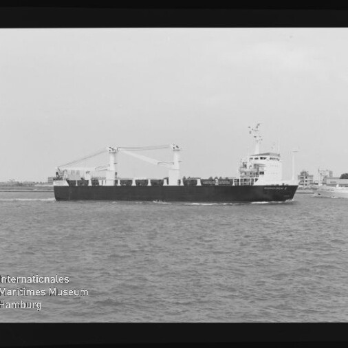 Schwarzweißaufnahme in Filmrahmen eines Frachtschiffs mit zwei Ladekränen, das auf ruhiger See an einer Küstenlinie vorbeifährt; unten links sichtbares Wasserzeichen des Internationalen Maritimen Museums Hamburg.