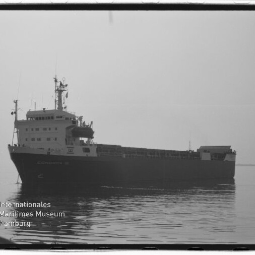 Schwarz-weiß-Aufnahme eines langen Frachtschiffs in ruhigem Wasser, seitliche Silhouette mit Brücke und Antennen, dezentes Spiegelbild auf der Wasseroberfläche und Wasserzeichen des Internationalen Maritimen Museums Hamburg unten links.