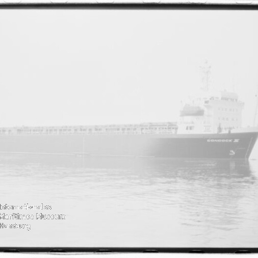 Schwarz-weiß-Foto eines großen Frachtschiffs, das in dichtem Nebel auf ruhigem Wasser treibt, mit leichter Spiegelung und sichtbarem Museumslogo unten links.