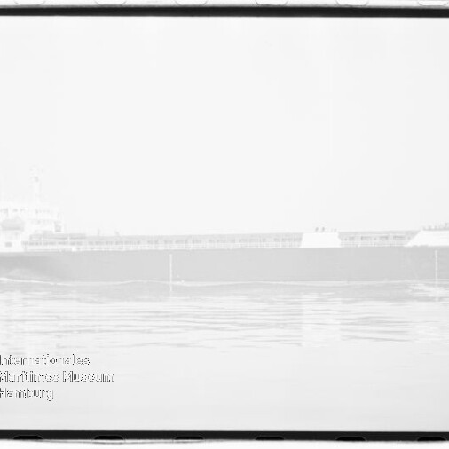 Schwarzweiß-Hochtonfoto eines langen Massengutfrachters in Seitenansicht auf ruhigem, spiegelndem Wasser, mit deutlichem Filmrand und Museumswasserzeichen unten links.