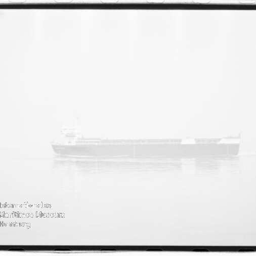 Schwarzweiß-Hochkontrastfoto eines Fracht-/Massengutfrachters, schwach sichtbar und fast überbelichtet, ruhig auf spiegelglattem Meer mit leichter Spiegelung, filmischer Rahmen und Stempel/Wasserzeichen unten links.
