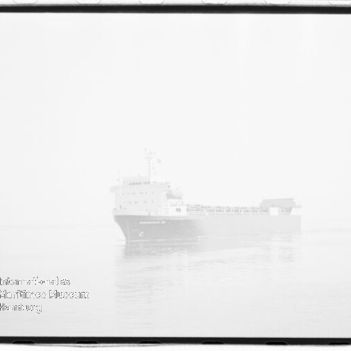Helles, im Nebel nahezu verschwommenes Frachtschiff auf ruhiger See mit Spiegelung im Wasser und einem Museumsstempel unten links.