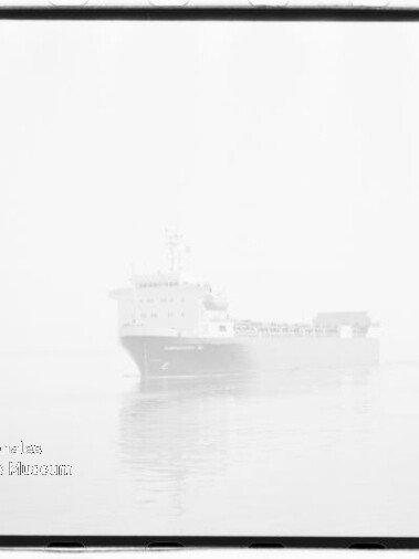 Großes Frachtschiff in dichtem Nebel auf ruhiger See mit schwacher Spiegelung im Wasser, sichtbarer Filmrahmenkante und Museumsstempel in der unteren linken Ecke.