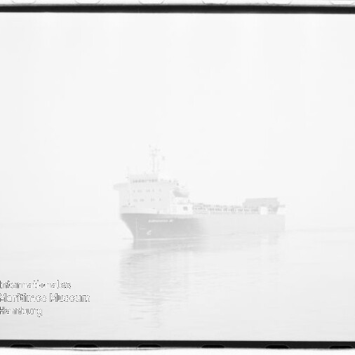 Großes Frachtschiff in dichtem Nebel auf ruhiger See mit schwacher Spiegelung im Wasser, sichtbarer Filmrahmenkante und Museumsstempel in der unteren linken Ecke.