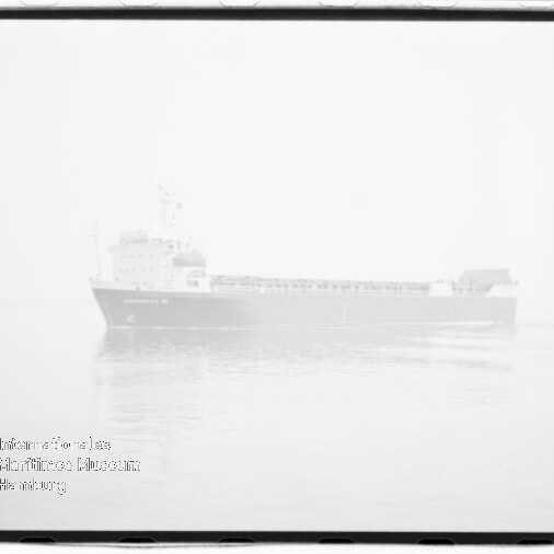 Schwarzweiß-Aufnahme eines großen Frachtschiffs in Seitenansicht auf ruhiger See vor hellem, überbelichtetem Himmel; schwaches Wasserzeichen des Internationalen Maritimen Museums Hamburg unten links und sichtbarer Filmrand.