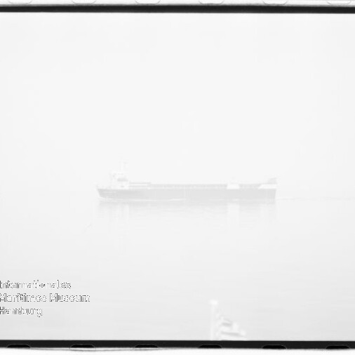 In dichtem Nebel zeichnet sich ein schwach sichtbares Frachtschiff auf ruhigem Wasser mit dezenter Spiegelung ab; das hochbelichtete Schwarzweißbild zeigt einen schwarzen Filmrand und ein Wasserzeichen unten links.