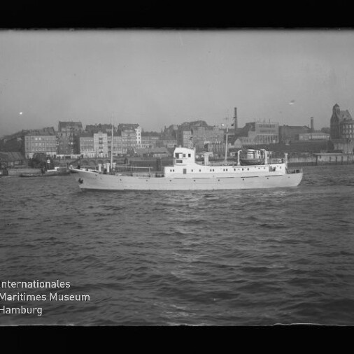 Weißes Motorpassagierschiff mit mittigem Aufbau fährt auf leicht bewegter Wasseroberfläche, im Hintergrund mehrstöckige Gebäude und Hafenbauten einer städtischen Uferzeile.