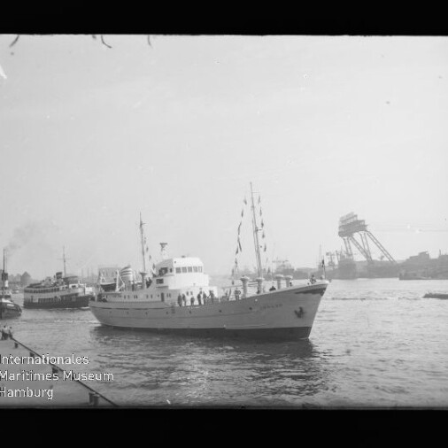 Schwarzweiß-Foto eines weißen Passagierboots mit Personen an Deck und Festtagswimpeln, das in einem Industriehafen zwischen weiteren Schiffen und großen Krananlagen fährt; unten links sichtbares Wasserzeichen des Internationalen Maritimen Museums Hamburg.