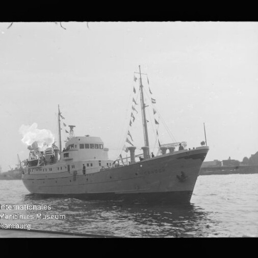 Mittelgroßes weißes Schiff mit beflaggtetem Mast und Rauch aus dem Schornstein fährt auf ruhigem Hafenwasser, im Hintergrund Uferbebauung; links unten Wasserzeichen des Internationalen Maritimen Museums Hamburg.