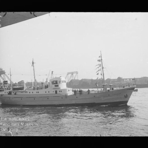Weißes Motor- bzw. Ausflugsschiff mit mehreren Masten und Flaggen fährt auf ruhigem Hafenwasser, im Hintergrund Hafenkräne und Hallen, unten links Wasserzeichen des Internationalen Maritimen Museums Hamburg.