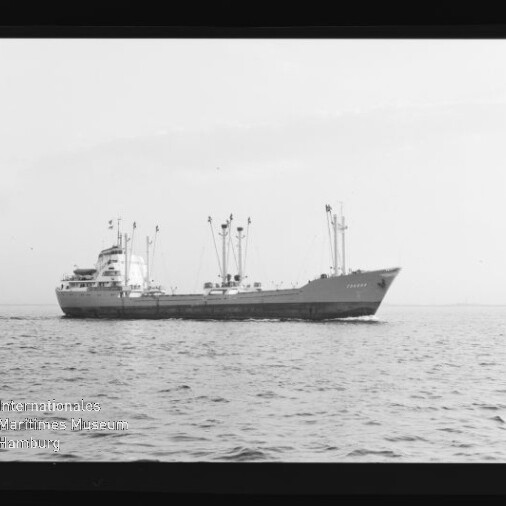 Schwarzweiß-Foto eines Frachtschiffs mit mehreren Ladekränen und Aufbauten, seitlich auf ruhiger See fahrend; Bildrand zeigt Filmstreifenmarkierungen und ein Wasserzeichen des Internationalen Maritimen Museums Hamburg.