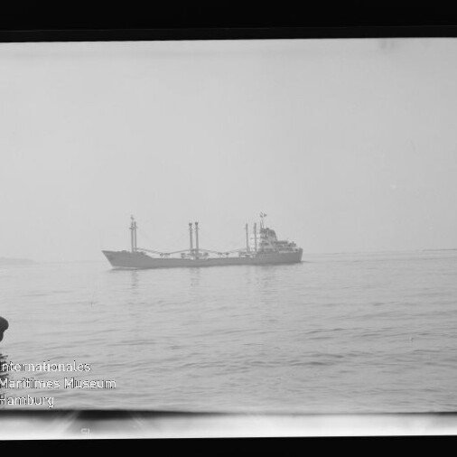 Schwarzweißaufnahme eines Frachtschiffs auf ruhiger See, im Vordergrund links zwei Männer auf einem kleinen Boot, mit schwarzem Bildrand und Wasserzeichen des Internationalen Maritimen Museums Hamburg unten links.