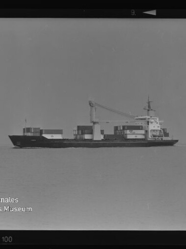 Schwarzweiß-Foto: Seitenansicht eines Containerschiffs mit aufgestapelten Containern und einem großen Ladekran auf ruhiger See vor klarer Horizontlinie.