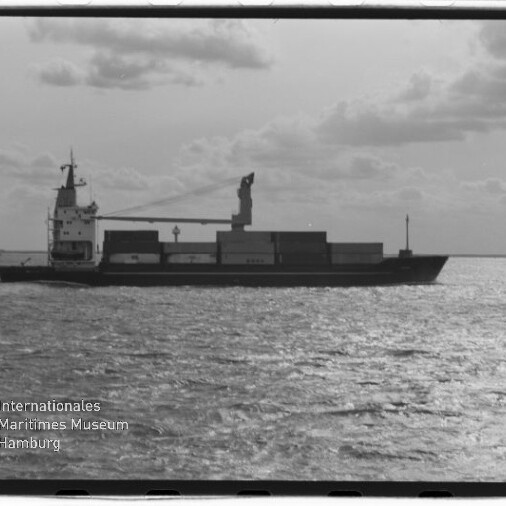 Schwarzweiß-Foto eines Frachtschiffs mit aufgestapelten Containern und einem Ladekran, das auf ruhiger See vor niedrigem Horizont und wolkigem Himmel fährt; unten links Logo und Schriftzug des Internationalen Maritimen Museums Hamburg.