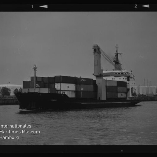 Schwarz-weiß-Aufnahme eines Containerschiffs im Hafen mit gestapelten Frachtcontainern und Bordkran, ruhige Wasserfläche im Vordergrund, sichtbarer Filmrand und Logo des Internationalen Maritimen Museums Hamburg links unten.