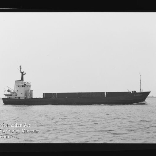 Schwarzweißaufnahme eines langen Frachtschiffs im Seitenprofil auf ruhiger See mit niedrigem Laderaum und Aufbauten am Heck; am Bildrand sichtbare Filmkanten mit Aufdruck AGFA APX 100 und Wasserzeichen des Internationalen Maritimen Museums Hamburg