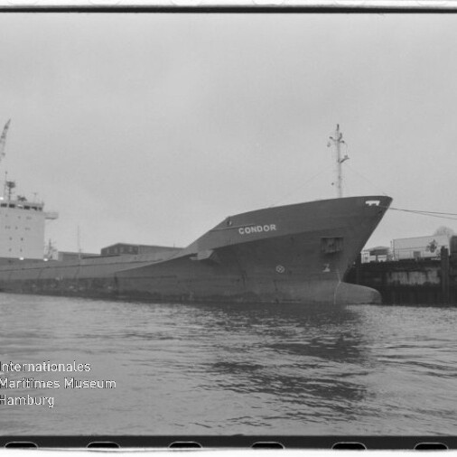 Schwarzweißaufnahme eines am Kai vertäuten Frachtschiffs mit dem Namen CONDOR, vor industriellem Hafen mit Kränen und Lagergebäuden, Wasser im Vordergrund und dem Logo des Internationalen Maritimen Museums Hamburg in der linken unteren Ecke.