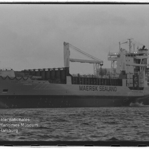 Schwarz-weiß-Seitenansicht des Frachtschiffs MAERSK SEALAND mit Ladekran und Aufbauten am Kai, Vordergrund Wasseroberfläche und links unten das Logo des Internationalen Maritimen Museums Hamburg.