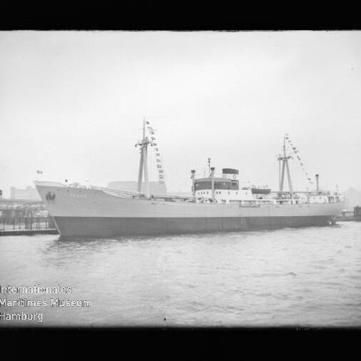 Schwarzweiß-Foto eines fest vertäuten Frachtschiffs mit beflaggten Masten am Kai, ruhiger Wasserfläche und sichtbarem Wasserzeichen des Internationalen Maritimen Museums Hamburg in der linken unteren Ecke.