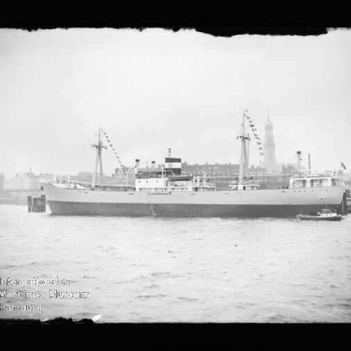 Schwarzweißaufnahme eines großen Schiffs mit zwei Masten und dekorativen Signalflaggen, am Kai liegend; im Vordergrund Wasser und ein kleines Lotsenboot, im Hintergrund eine Stadtsilhouette mit hohem Kirchturm; unten links Wasserzeichen des Internationalen Maritimen Museums Hamburg.