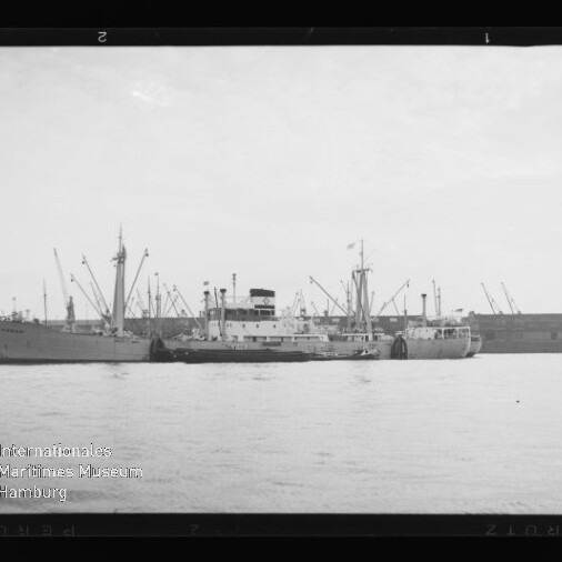 Schwarzweiß-Foto eines Frachtschiffs, das dicht begleitet von einem Schlepper parallel am Hafen liegt, vor einer Kaizeile mit zahlreichen Kränen; unten links sichtbar das Wasserzeichen des Internationalen Maritimen Museums Hamburg.