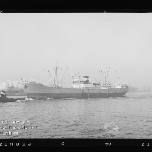 Schwarzweiß-Foto eines Frachtschiffs mit Ladekränen, begleitet von einem Schlepper im Hafen bei Nebel; im Vordergrund Wasser und links unten das Logo des Internationalen Maritimen Museums Hamburg.