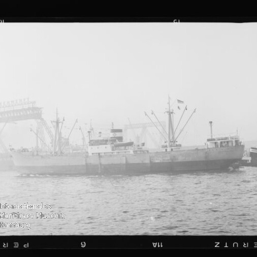 Seitlich abgebildetes Frachtschiff an einem Industriehafen mit mehreren Ladekränen im Hintergrund, ruhige See im Vordergrund und leicht nebliger Dunst.