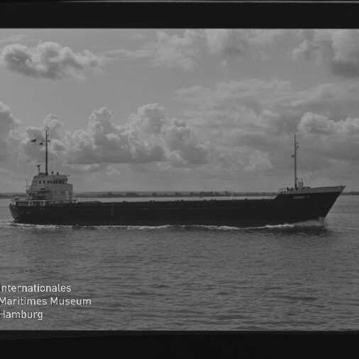 Schwarzweißaufnahme eines länglichen Frachtschiffs in Seitenansicht, das ruhig über das Wasser fährt vor einem wolkenbedeckten Himmel; unten links sichtbar das Wasserzeichen Internationales Maritimes Museum Hamburg.
