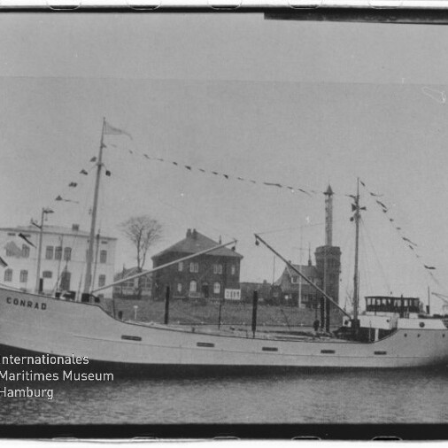 Schwarz-weiß-Foto eines Frachtschiffs mit dem Namen CONRAD am Bug, das mit Wimpelketten geschmückt am Ufer liegt; im Hintergrund Häuser und ein hoher Turm, links unten Wasserzeichen des Internationalen Maritimen Museums Hamburg.