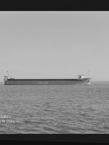 Schwarzweißaufnahme eines langen Frachtschiffs mit lesbarer Rumpfbeschriftung, das ruhig nahe dem Horizont auf ruhiger See fährt; Filmrand und Museumswasserzeichen sind sichtbar.