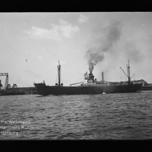 Schwarz-weiß-Foto eines dampfbetriebenen Frachtschiffs mit rauchendem Schornstein, das auf dem Wasser an einer Hafenkaianlage mit Kränen im Hintergrund vorbeifährt.