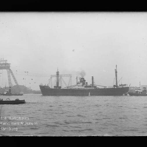 Schwarzweiß-Foto eines großen Dampfschiffs mittig auf dem Wasser, mit Rauch aus dem Schornstein, im Vordergrund wellige Wasserfläche und links ein kleines Boot, im Hintergrund Hafenkräne und Industrieanlagen; im unteren linken Bildbereich das Logo des Internationalen Maritimen Museums Hamburg.