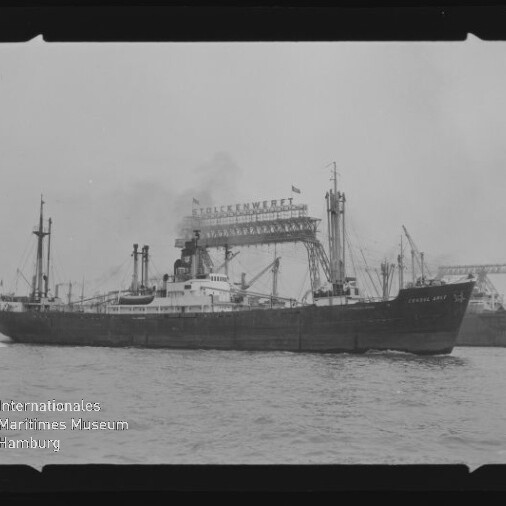 Schwarzweißaufnahme eines Frachtschiffs, das auf dem Wasser an einer Werft mit großen Auslegerkränen und dem Schriftzug „Stülckenwerft“ vorbeifährt; unten links ist ein Museumswasserzeichen zu sehen und der Filmrand bleibt erkennbar.