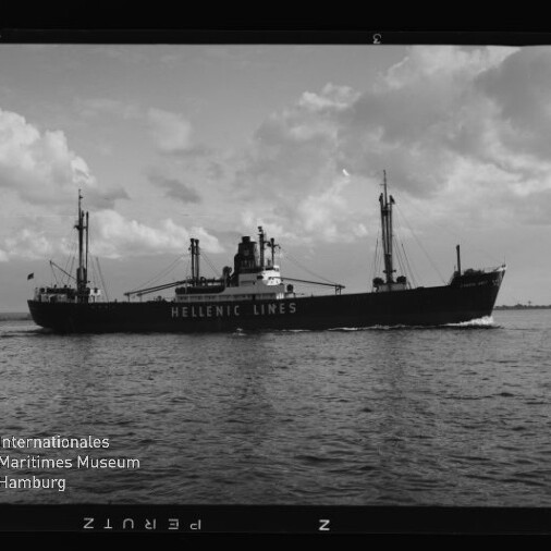 Schwarzweißaufnahme eines Frachtschiffs der Reederei Hellenic Lines mit sichtbarem Schriftzug auf dem Rumpf, Ladebäumen und Schornstein, das bei ruhiger See unter teilweise bewölktem Himmel von links nach rechts fährt; unten links das Logo des Internationalen Maritimen Museums Hamburg.