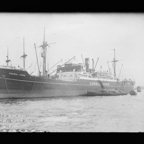 Schwarzweiß-Foto eines großen Frachtschiffs mit dem Namen CONSUL HORN, das am Kai liegt; mehrere Masten und ein Schornstein sind sichtbar, kleinere Boote liegen am Rumpf und Wasserzeichen des Internationalen Maritimen Museums Hamburg ist unten links zu sehen.