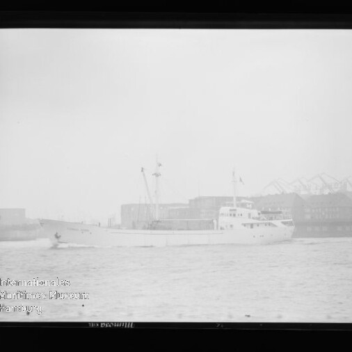 Weißes Frachtschiff mit Aufbauten und zwei Masten in einem nebligen Industriehafen; im Hintergrund Lagergebäude und mehrere Hafenkräne, im Vordergrund wellige Wasseroberfläche und unten links ein Museumsstempel.