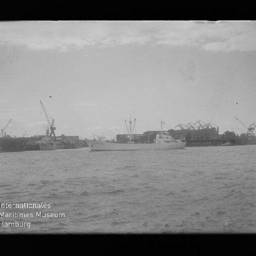Schwarzweißaufnahme eines mittleren weißen Schiffes auf dem Wasser vor einer Werft mit Kränen und Hallen, mit Museumslogo unten links.