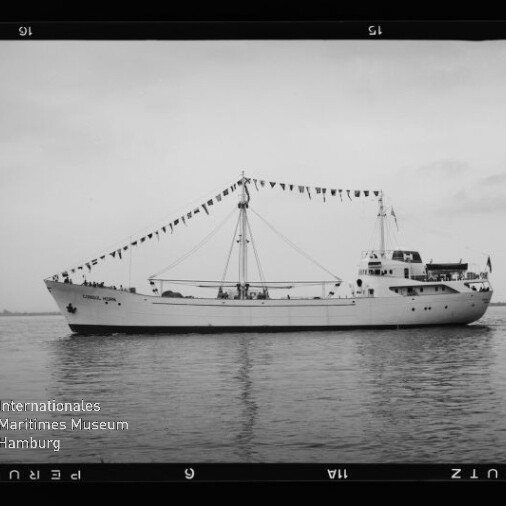 Seitenansicht eines weißen Schiffes mit zentralem Mast, an dem eine Girlande aus Signalfahnen hängt; das Boot liegt auf ruhigem Wasser vor flachem Horizont, im Foto sind Filmrand und ein Museumslogo zu sehen.