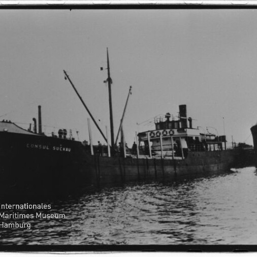 Schwarzweißaufnahme eines im Hafen liegenden Frachters mit Ladebäumen und Schornstein, auf dessen Bug der Name 'KONSUL SUCKAU' zu lesen ist; im Vordergrund Wasser, unten links das Wasserzeichen des Internationalen Maritimen Museums Hamburg.