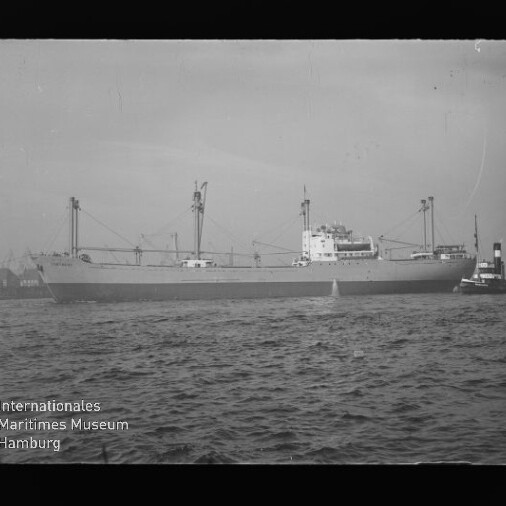 Schwarzweißfoto eines großen Frachtschiffs mit Ladekränen und Aufbauten, das auf dem Wasser liegt und am Heck von einem Schlepper begleitet wird; unten links ist das Wasserzeichen des Internationalen Maritimen Museums Hamburg.