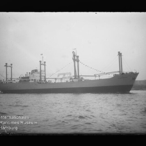 Schwarzweiß-Aufnahme eines Frachtschiffs in Seitenansicht auf ruhiger See mit mehreren Ladebäumen und Girlanden an den Masten; unten links ist das Logo des Internationalen Maritimen Museums Hamburg sichtbar.