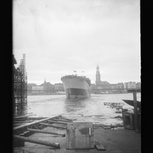 Großes Passagierschiff fährt in einen Hafen; im Vordergrund Kaianlage mit Holzbohlen und einer Kiste, im Hintergrund die Hamburger Stadtsilhouette mit dem Turm der St.-Michaelis-Kirche.